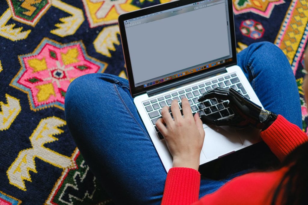 somebody sat crossed legged on the floor using a laptop. Their right hand is a black prosthetic