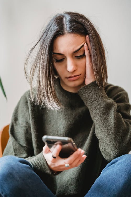 a young woman with long brown hair, sat on a sofa looking stressed at her phone, with her hand across her face