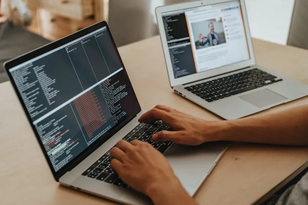 someone typing on one laptop with coding on the screen, next to another with someone talking over a online call