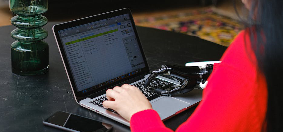 a woman wearing a red top and with a right prosthetic hand sat at a dining able using a laptop