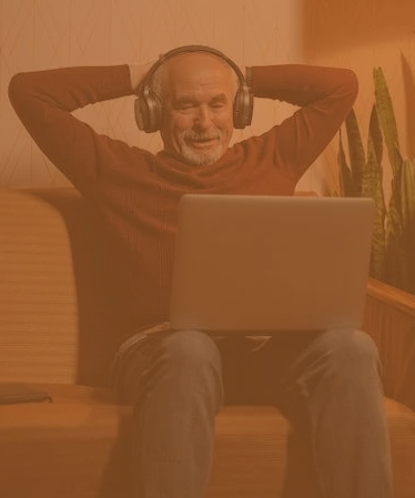 an older man sat with his hands behind his head, wearing headphones, and enjoying watching something on his laptop