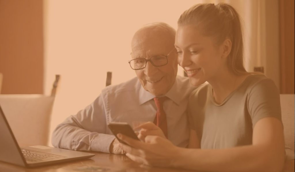 a young lady sat with an elderly male, at a dining room table showing him how to use his mobile phone and laptop