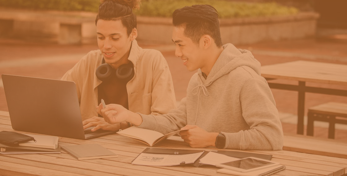 two students sat laughing on a bench, next to a laptop