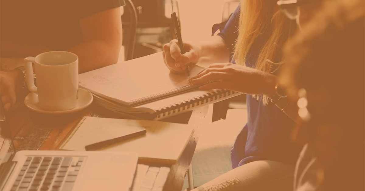 a woman sat taking notes in a pad, whilst sat at a laptop, next to a cup of coffee