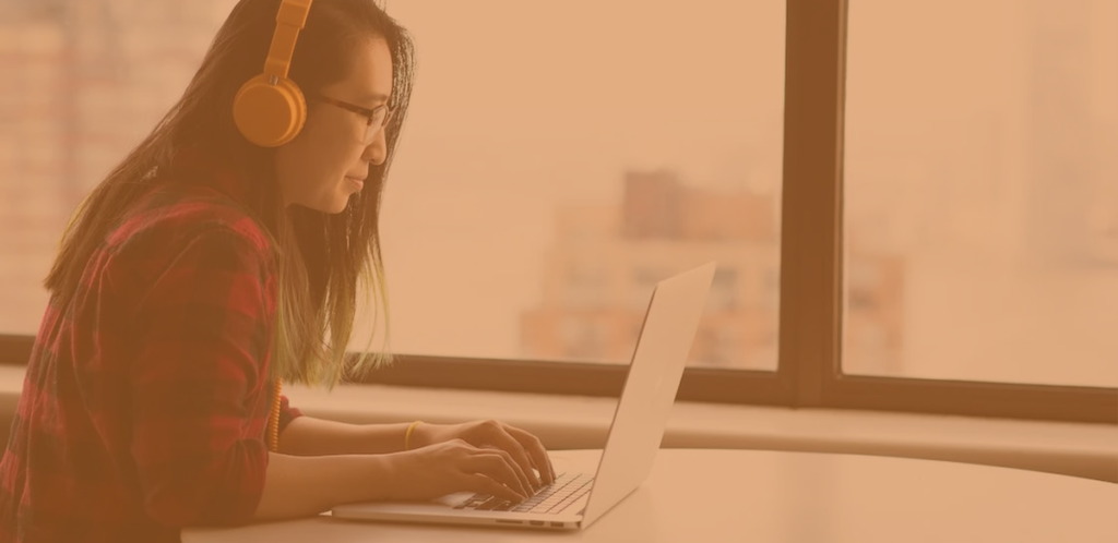 a young woman with long black hair, sat working on a laptop, wearing glasses and an orange pair of headphones