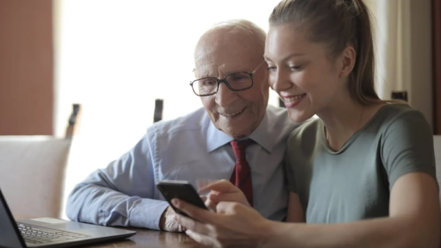 an old man sat with a young woman who is showing him how to use a mobile phone and laptop