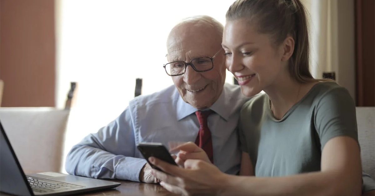 an old man sat with a young woman who is showing him how to use a mobile phone and laptop