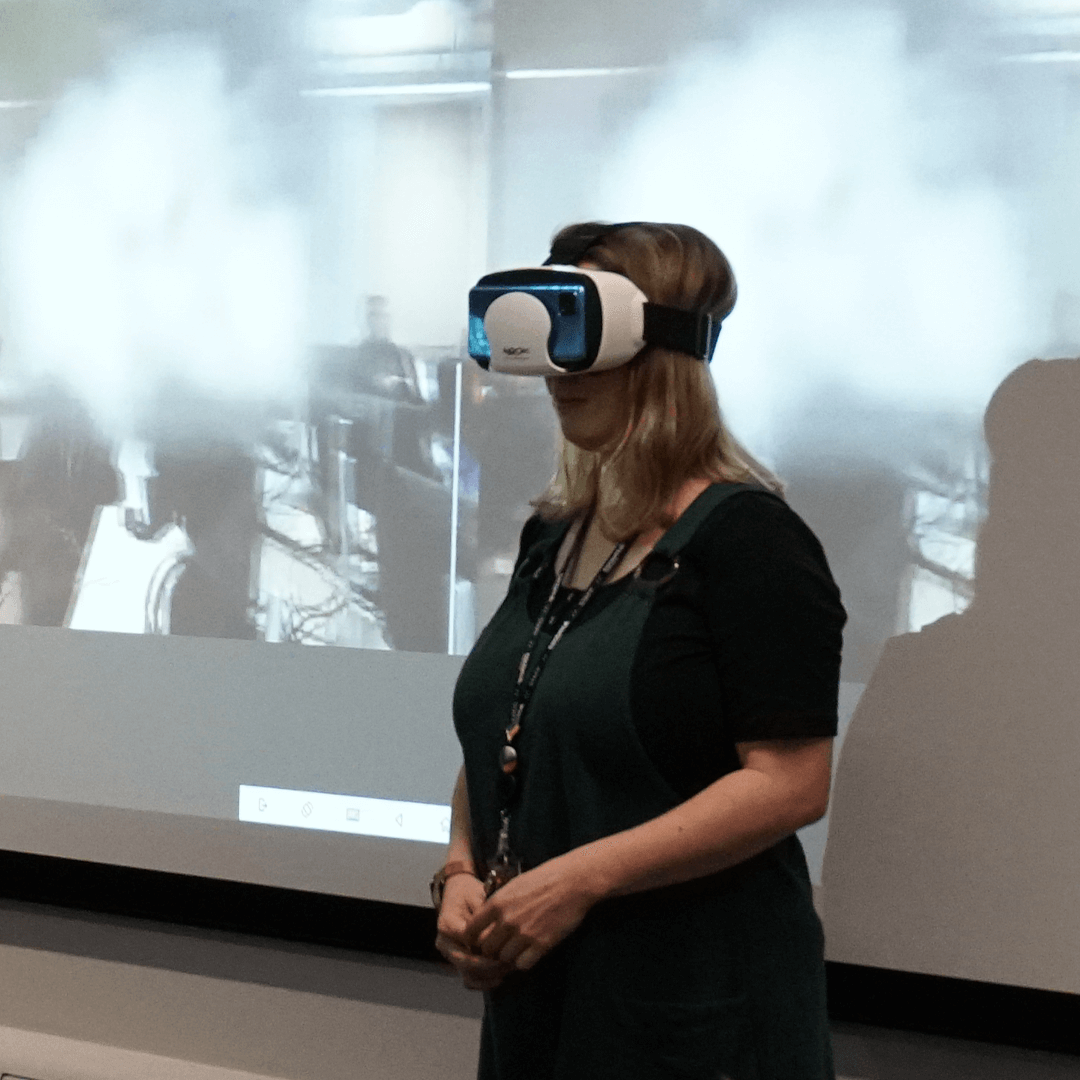 A university lecturer stood wearing a VR headset, in front of a projector that displays what she is seeing through VR