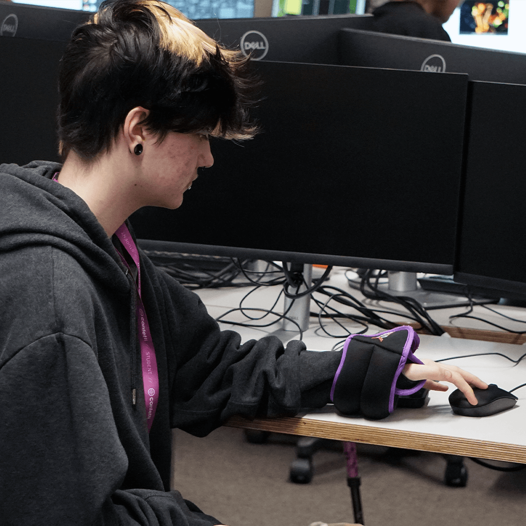 A student sat at a desk, trying to use a mouse, whilst wearing weights on his wrists