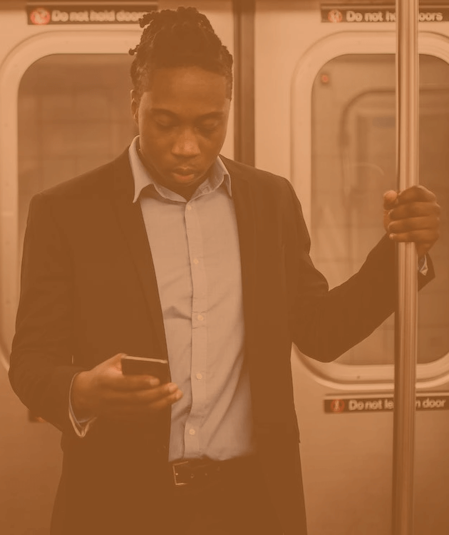 a Black man stood holding onto a handrail on a train, whilst watching a video on his mobile phone