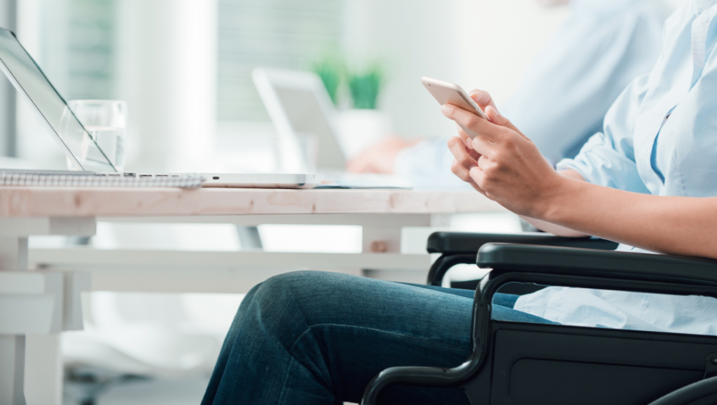 a wheelchair user, next to a desk with a laptop on whilst using a mobile phone
