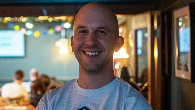 Tom Miller smiling, wearing a t-shirt with a purple accessibility icon in the middle within a hexagon, at an Accessibility Nottingham event