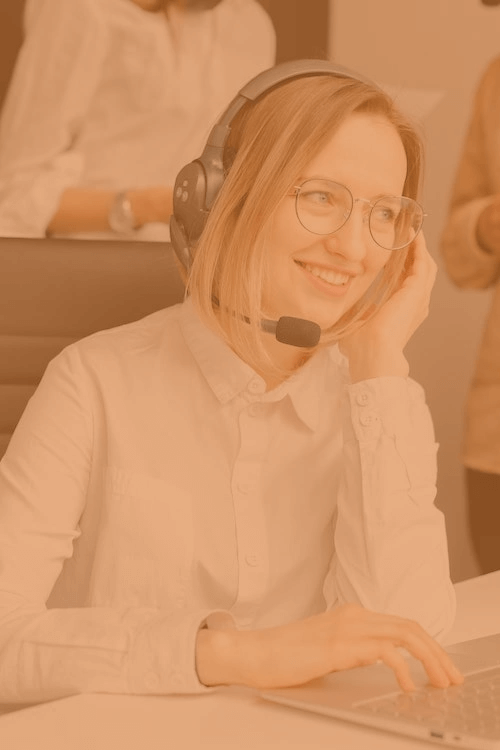 a young woman with blonde hair, sat smiling at a work station, whilst wearing a headset and typing on a laptop