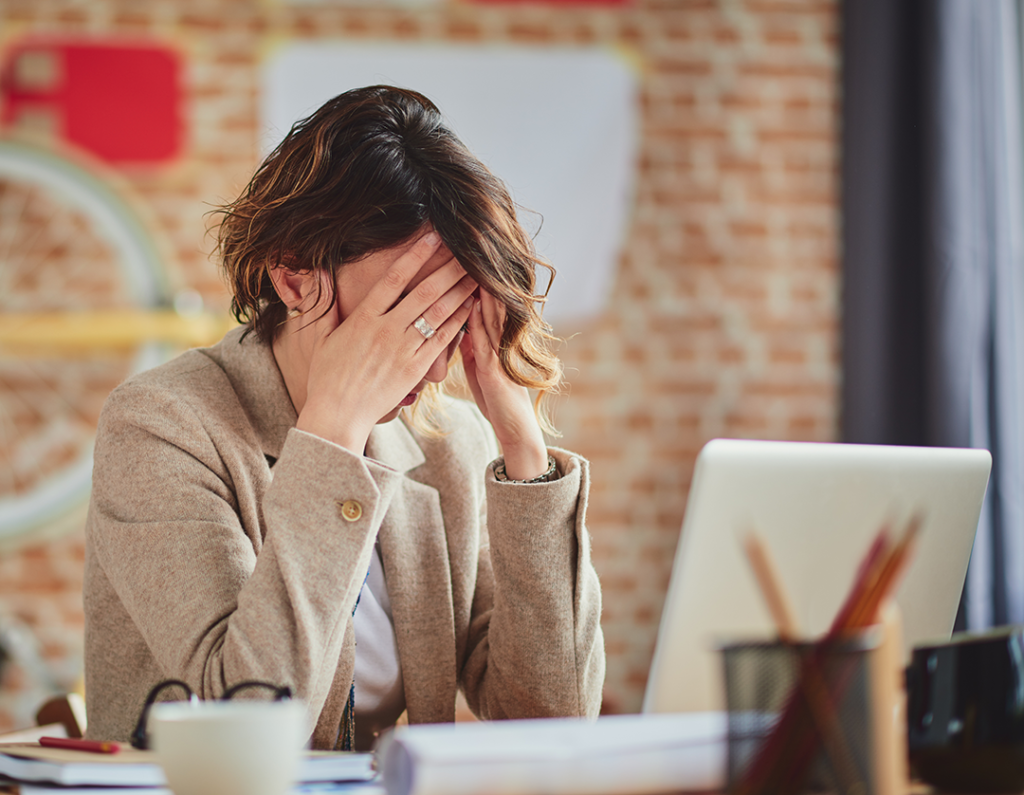 a woman sat at a desk next to a laptop with her head in her hands in distress