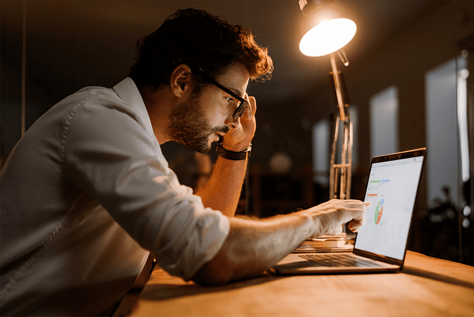 a man sat in front of his laptop adjusting his glasses, whilst trying to read a graph on his laptop screen