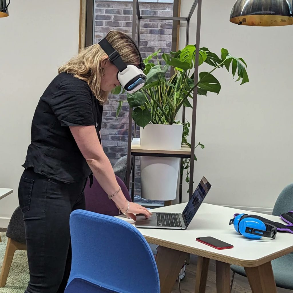 a woman with blonde hair is stood wearing a VR headset, looking down at a laptop screen and trying to navigate through a website with the touchpad