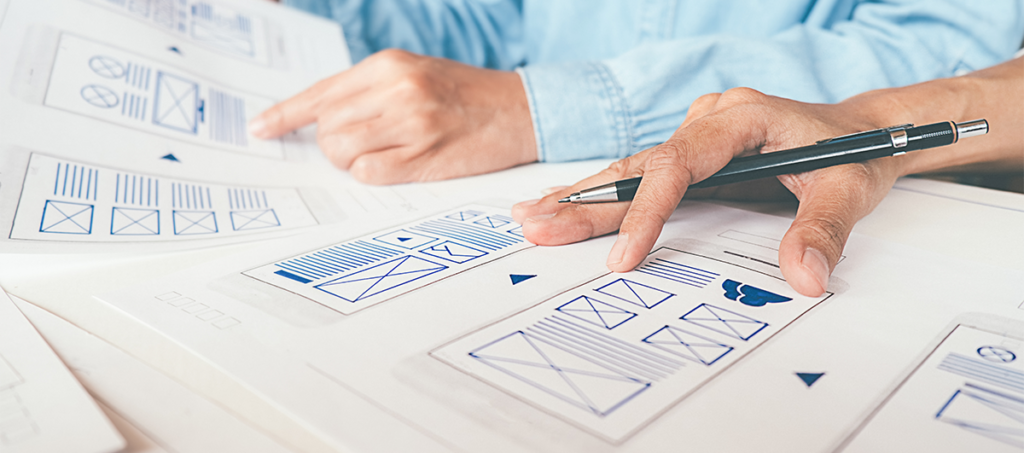 two men sat at a table sketching out their website plans, drawing boxes where content will sit on four different pieces of paper.