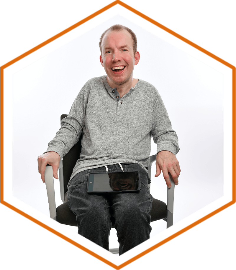 Lee Ridley, AKA Lost Voice Guy, sat in a chair smiling with a black tablet on his knee, against a white backdrop. Lee is wearing a grey jumper and black jeans.