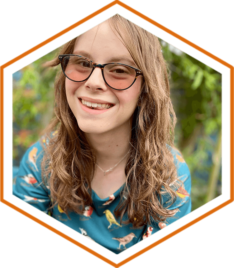 Chloe Tear in a garden wearing brown glasses, with a huge smile on her face. She is wearing a blue top which has different types of colourful birds all over it.