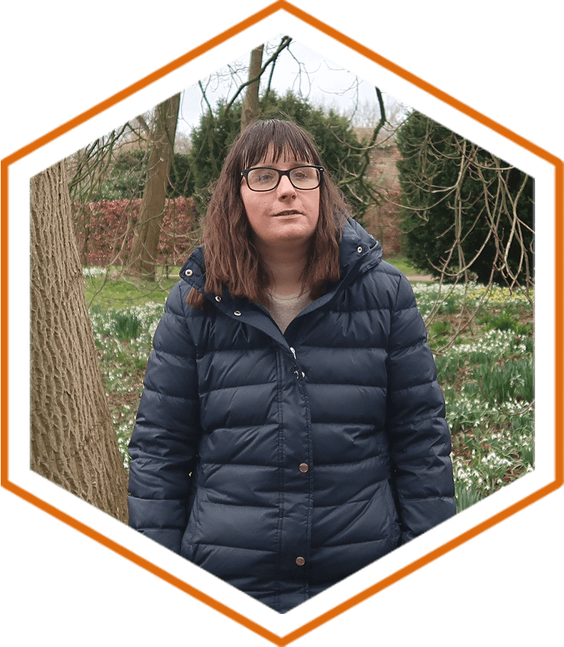 Holly stood on a path with a mass of white snowdrops and brown trees in the distance behind her. She's wearing a blue coat and black and grey trousers