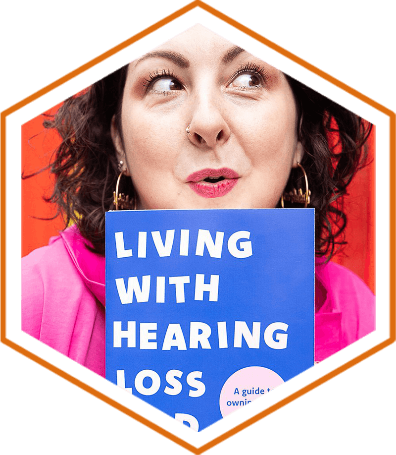 A head and shoulder shot of Samantha Baines who has a black curly bob and brown eyes. She stands in front of a red background wearing a pink top and gold hoop earrings. She holds up her book to her chin and looks off to the side cheekily. The title of the book is Living With Hearing Loss And Deafness, written in pale pink on a blue book cover.