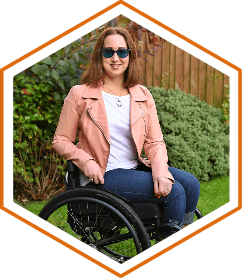 Tania Dutton is in a wheelchair in a garden surrounded by shrubbery. Tania has brown shoulder length hair and is smiling, whilst wearing green tinted sunglasses, a white top, jeans and rose-coloured leather jacket