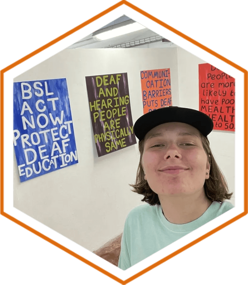 Zoe Milner in a mint green t-shirt and cap, stood smiling in front of two pieces of her artwork which has large typography which reads ‘BSL act now protect deaf education’ and ‘Deaf and hearing people are physically the same’