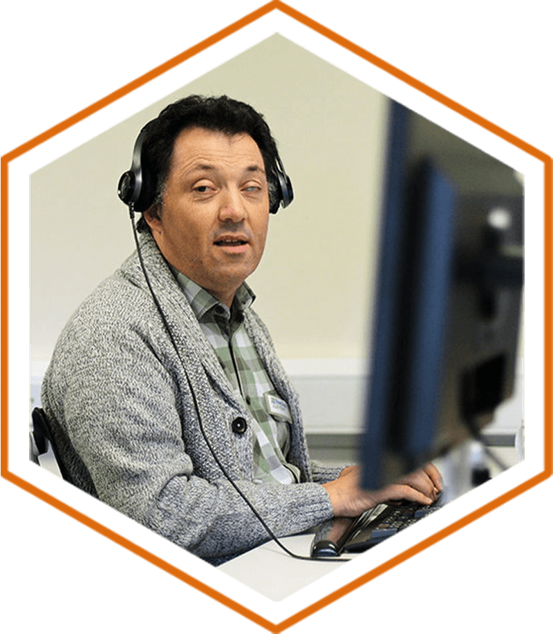 Alan Sleat, who has short black hair and is wearing a pale green shirt and grey cardigan, is sat at his desk using a desktop computer and wearing a black headset