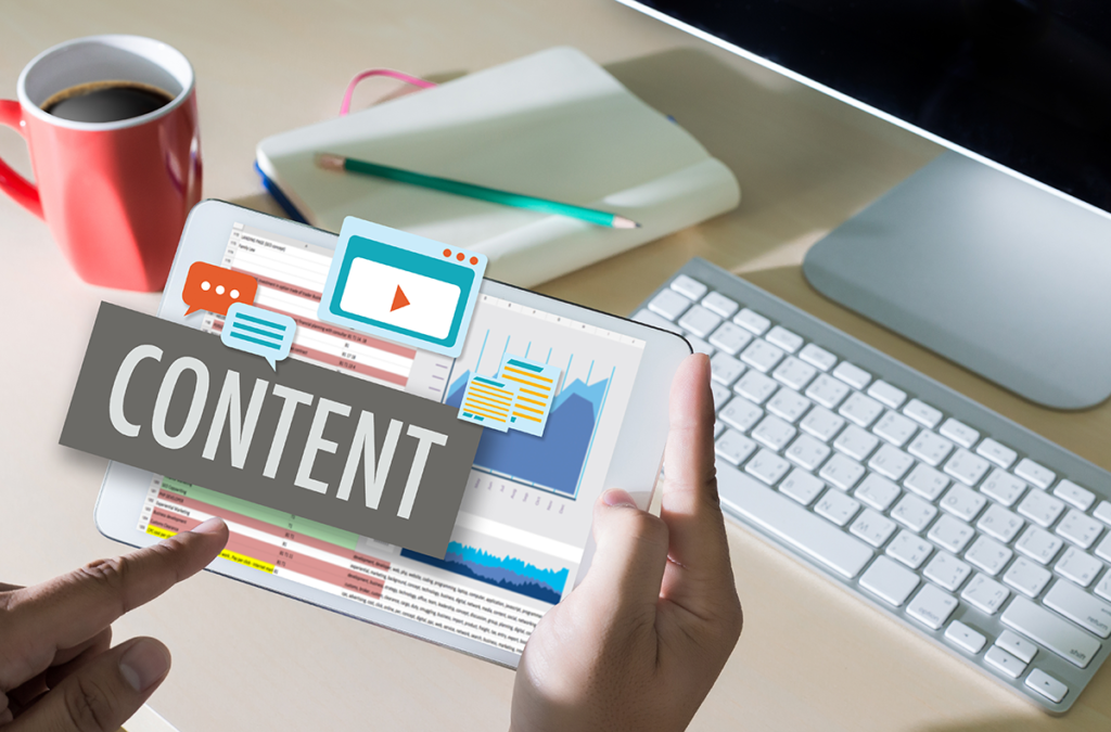 someone's hands holding a tablet, coming out above the screen is the word "content" and icons such as documents, videos, comments, and analytics. This is in front of a desk with a computer and keyboard in the background.