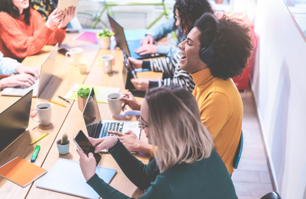 a communications team sat around a shared desk. All of them are working hard on their laptops and mobile devices. In the centre of the image is a Black man, who is wearing a bright yellow top and a headset, who has his head back laughing.