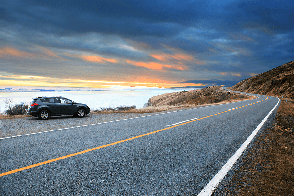 A dark blue 4x4 car is sat in a lay-by next to a winding rural road at sunrise. The car is looking out over a bright turquoise-coloured lake with white clouds that low down, they are virtually on top of the lake itself.