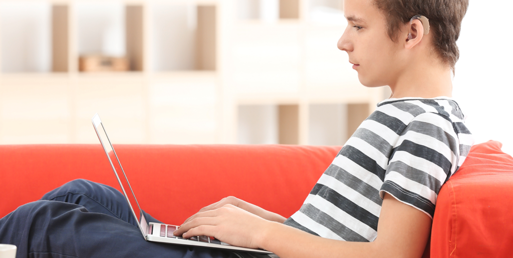 young boy is sat on a red sofa in his living room watching a video on a laptop. He has short brown curly hair and is wearing a striped t-shirt, blue jeans, and has a hearing aid in his right ear.