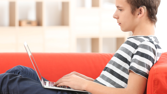 young boy is sat on a red sofa in his living room watching a video on a laptop. He has short brown curly hair and is wearing a striped t-shirt, blue jeans, and has a hearing aid in his right ear.