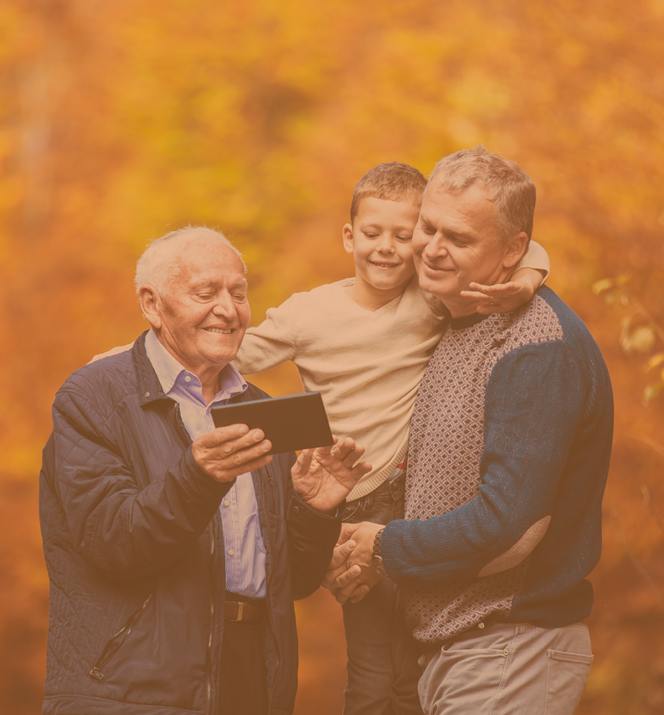 an old man stood surrounded by autumnal trees is showing his son and grandson a website on his tablet