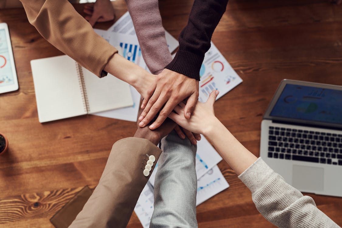 5 people from different races are all piling their hands on top of each other in a circle to do a upwards high five. This is above an office desk with a laptop and reports under them.