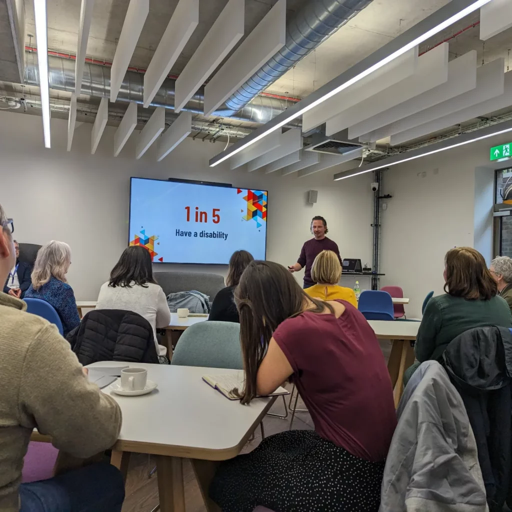 HeX's Creative Director James Hall is presenting a training session to a large room full of people. On the screen behind him it reads that 1 in 5 people have a disability