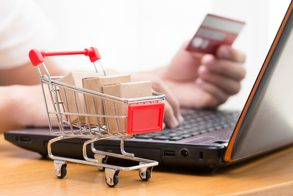 a tiny shopping trolley has three parcels inside wrapped in brown paper. The trolley is next to a large laptop and someone using their credit card to make an online purchase.