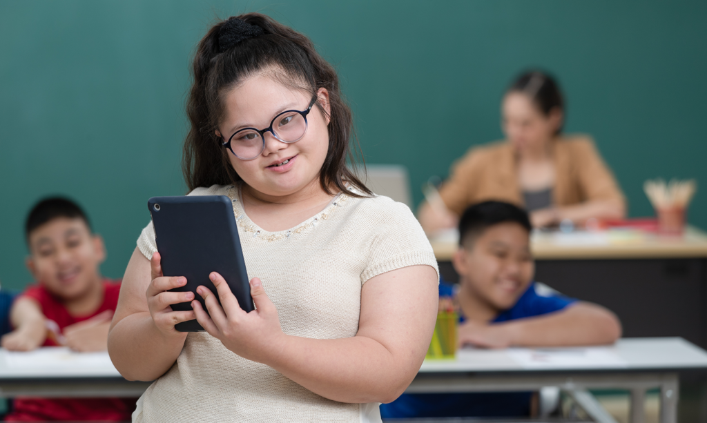 a young Asian girl who has Downs Syndrome is stood in front of a class full of students. She is smiling and tilting her head to one side whilst wearing glasses and holding a tablet