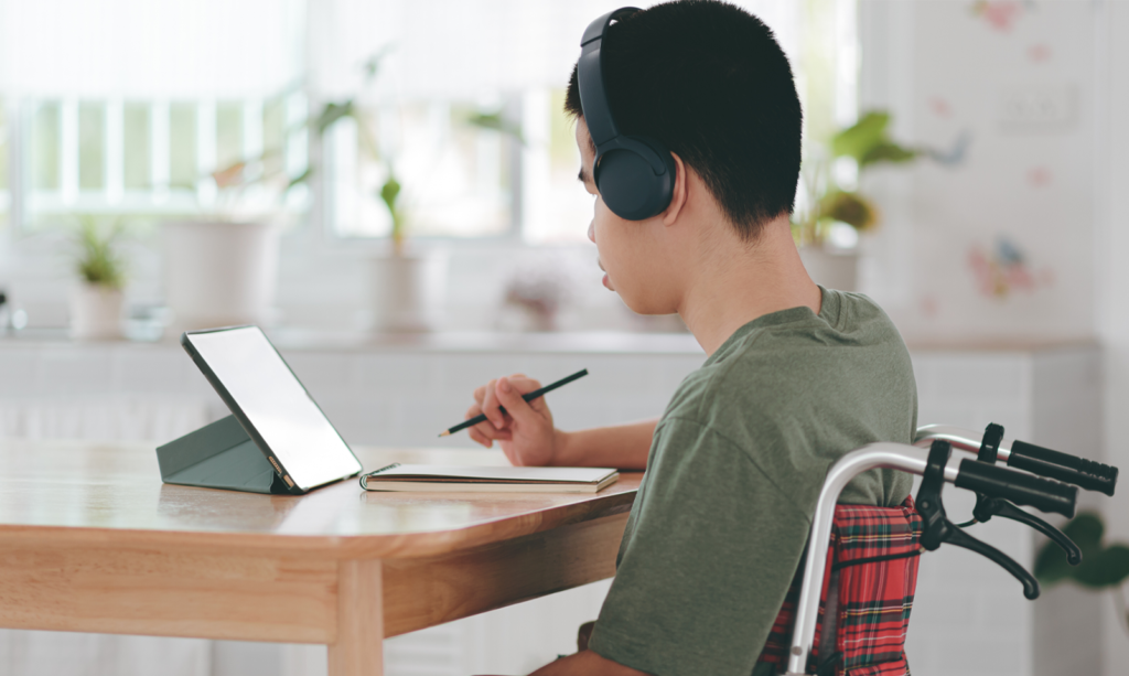 a young boy is in his wheelchair at his kitchen table. He is sat wearing a green t-shirt and black headphones, whilst making notes from accessing a tablet