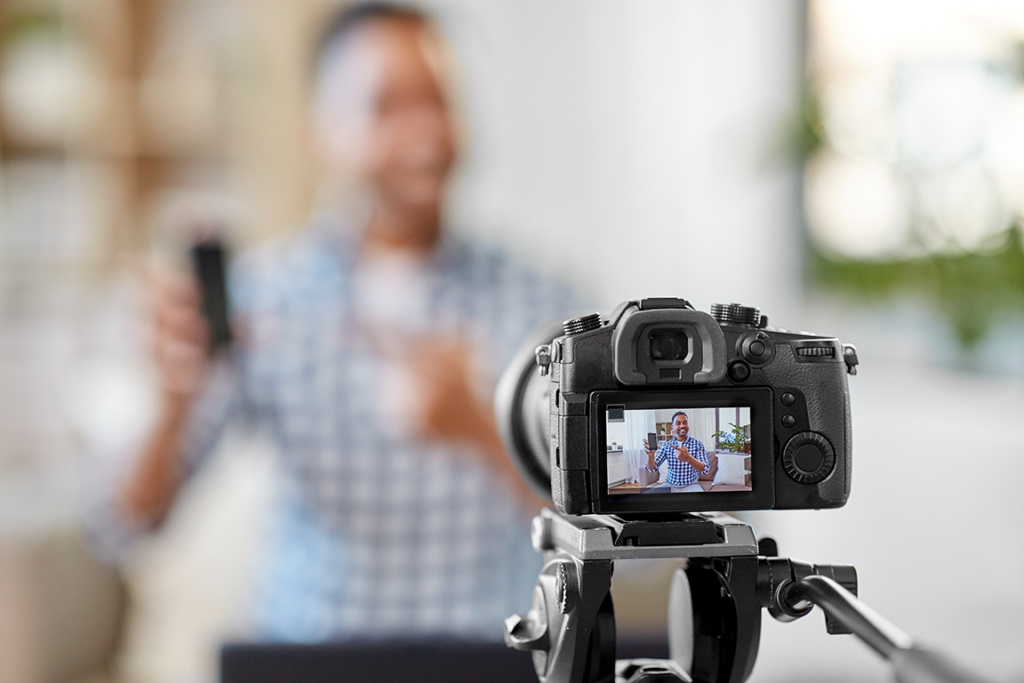 a man wearing a checked shirt is sat in front of a DSLR camera. He is being filmed whilst selling a mobile phone.