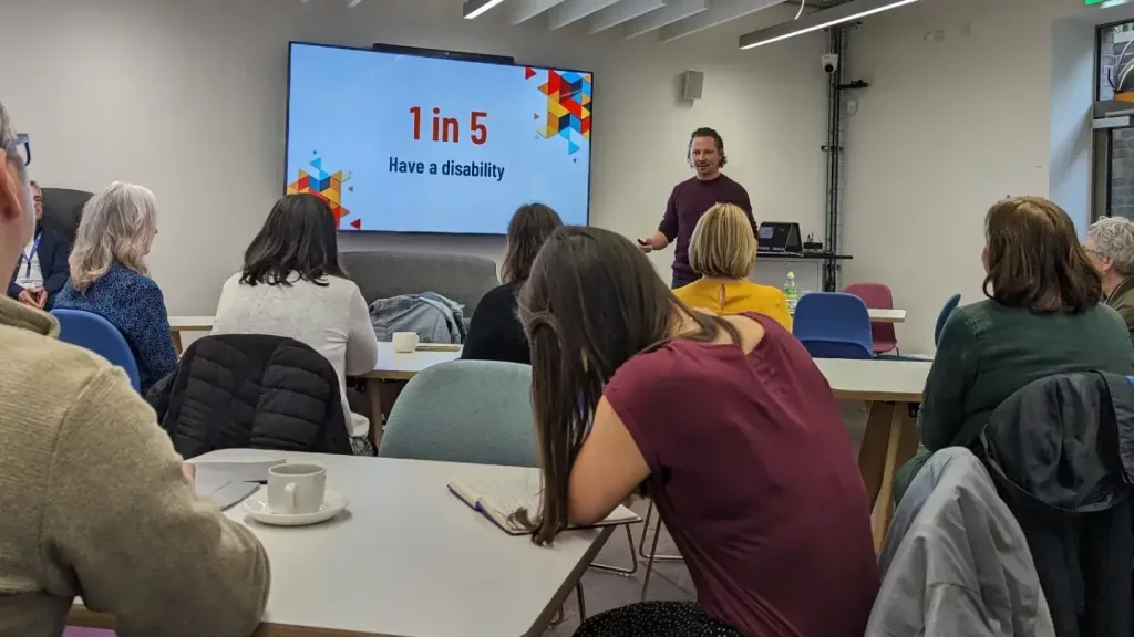 HeX's Creative Director James Hall is presenting a training session to a large room full of people. On the screen behind him it reads that 1 in 5 people have a disability