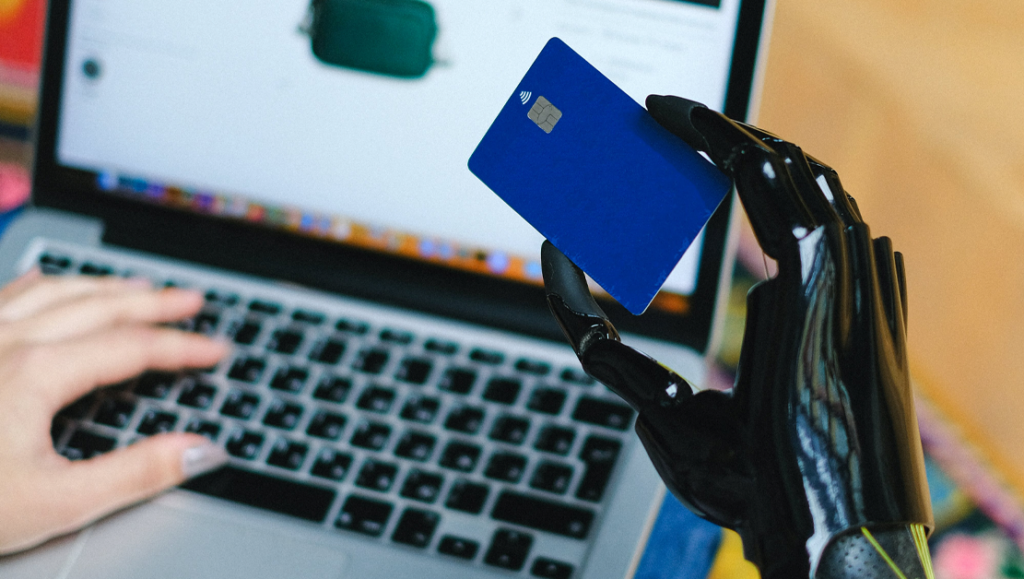 someone is typing on a laptop with their left hand and holding a blue credit card with a prosthetic hand with their other.