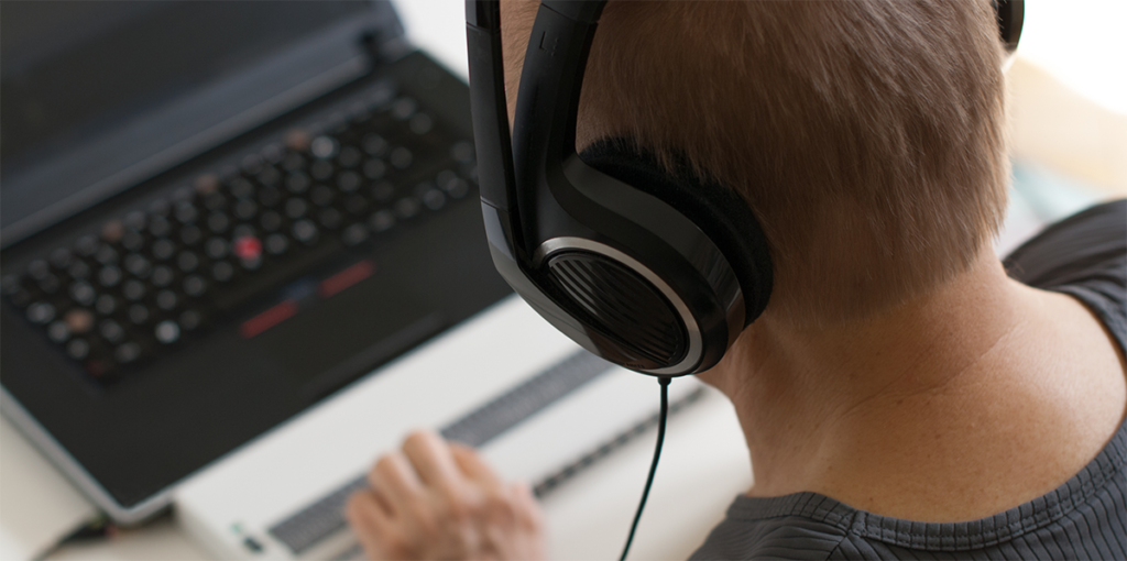 the back of a man with short brown hair, who is sat wearing black headphones and typing on a Braille keyboard which is connected to a black laptop
