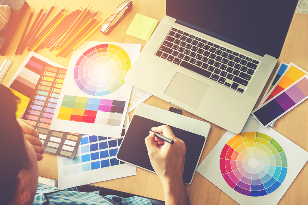 a graphic designer is sat at their desk in front of a laptop and surrounded by colour wheels and pallets. They are sketching on a drawing tablet.