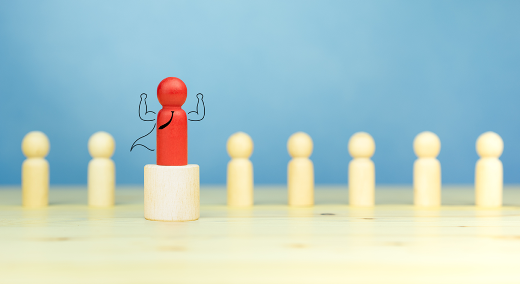 7 beige wooden pegs in the shape of people are stood in a row, in the centre is a red peg which has a tie, cape and arms reaching to the sky hand-drawn on