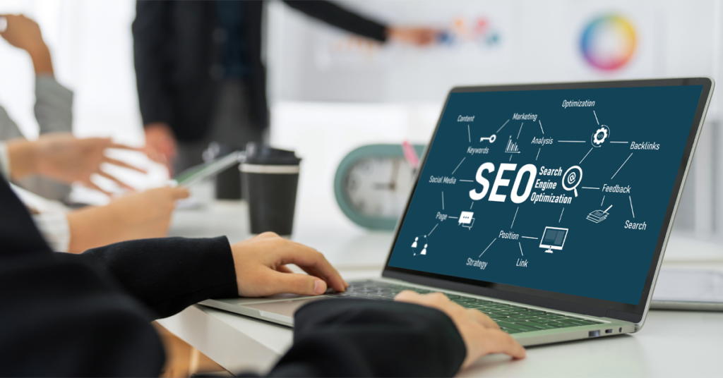SEO and website accessibility - someone typing into a laptop on an office desk next to a cup of coffee, the screen has the wording SEO across it. In the background someone is stood pointing to an array of colourful graphs.