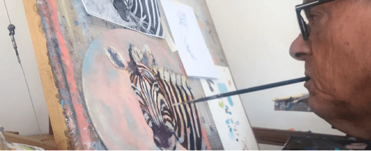 Mouth and Foot Painting Artist Rob Trent is holding a paintbrush in his mouth whilst painting a colourful picture of a zebra on canvas.