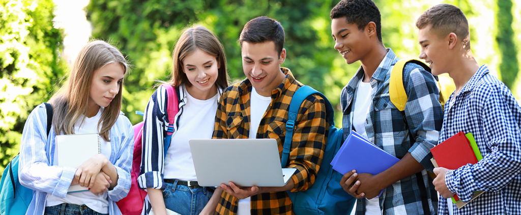 A group of college students are stood together outdoors all looking towards a university website on one of the students laptop