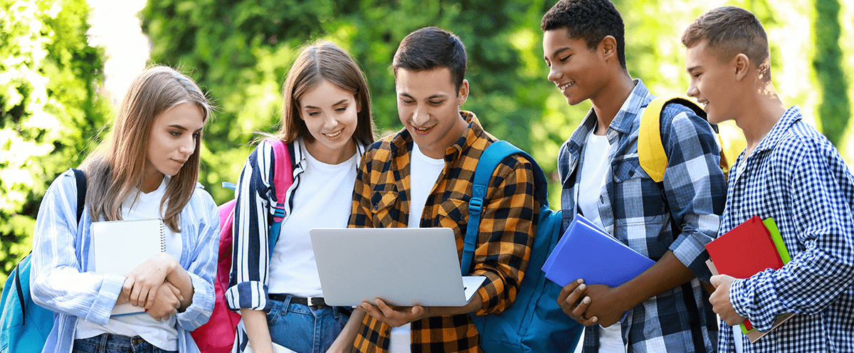 A group of college students are stood together outdoors all looking towards a university website on one of the students laptop