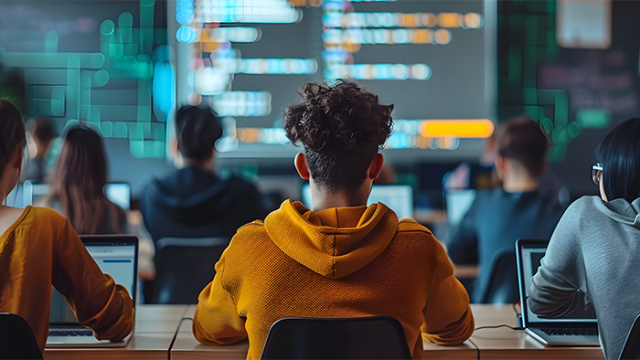 A class of students are all sitting in front of rows of laptops facing a white board which is displaying code